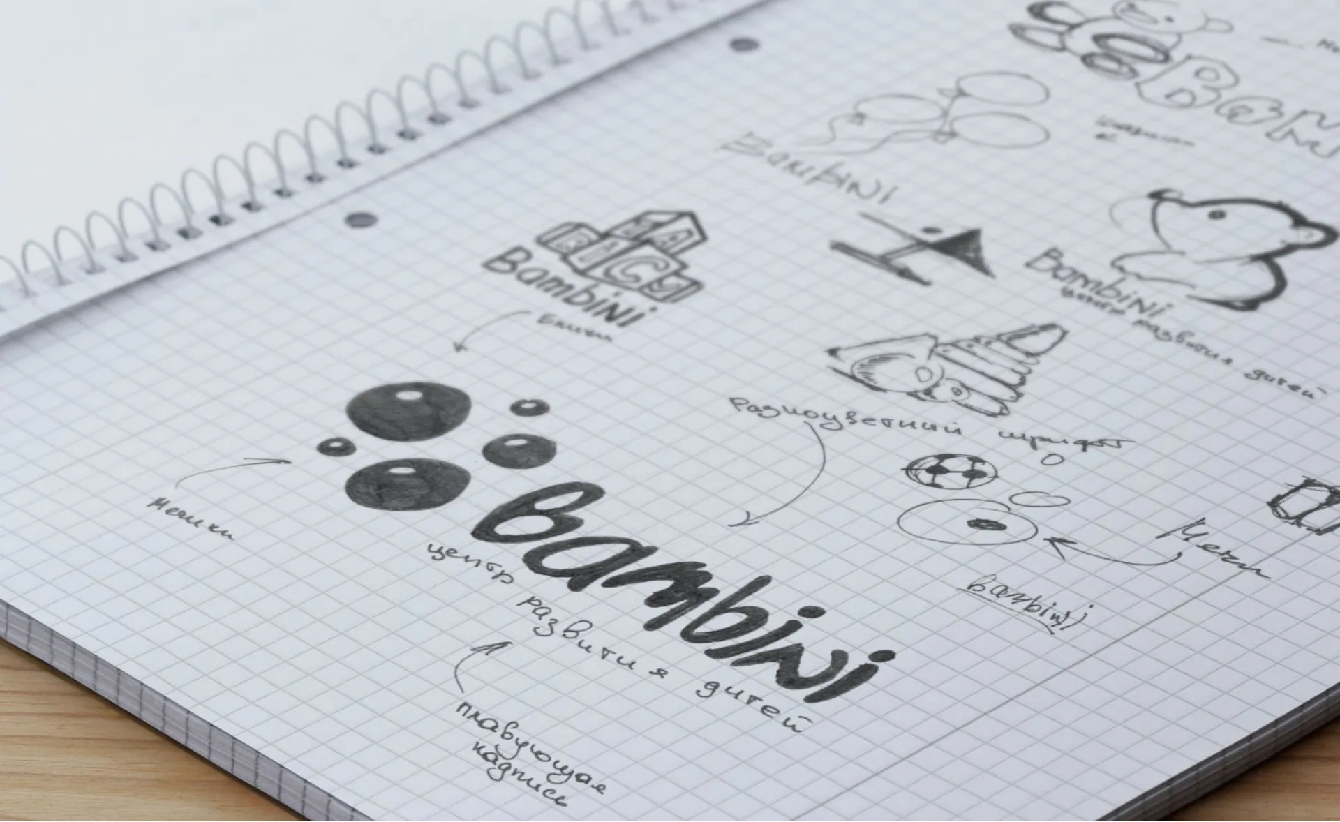 Эскизы логотипа Bambini в блокноте в клетку: кубики, мишка, шарики